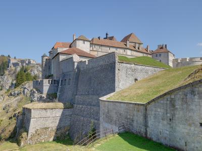 A large stone fortress with terracotta-tiled roofs, grassy ramparts, and defensive walls built into a rocky hillside under a clear blue sky.