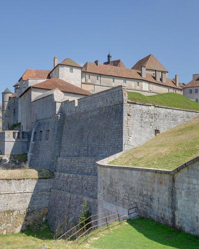 A large stone fortress with terracotta-tiled roofs, grassy ramparts, and defensive walls built into a rocky hillside under a clear blue sky.