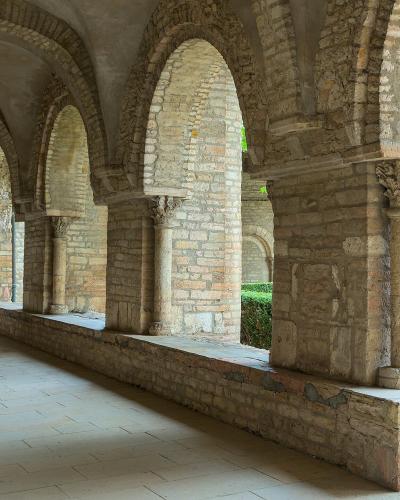 The image shows a stone cloister with rounded arches, columns with carved capitals, and a paved walkway adjacent to a garden area.