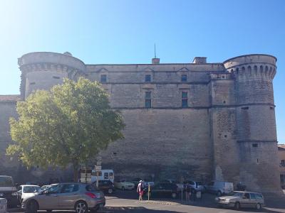 A large medieval stone fortress with two prominent round towers and a high curtain wall, situated behind a parking lot with cars and a tree in the foreground.