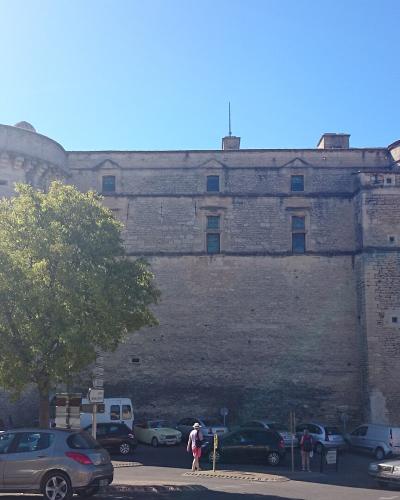 A large medieval stone fortress with two prominent round towers and a high curtain wall, situated behind a parking lot with cars and a tree in the foreground.