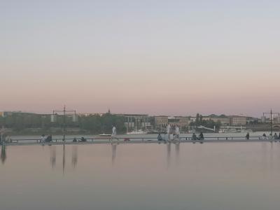 A wide, shallow reflecting pool at dusk with people walking and sitting along its edge, and a riverfront cityscape with buildings and boats in the background.