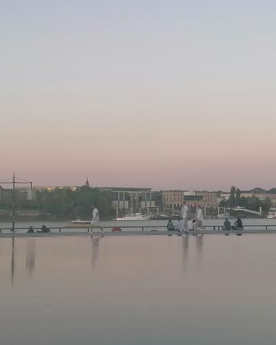 A wide, shallow reflecting pool at dusk with people walking and sitting along its edge, and a riverfront cityscape with buildings and boats in the background.