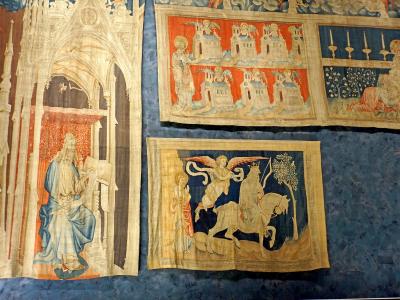The image shows several large, medieval tapestries with intricate religious and apocalyptic scenes, displayed on a dark blue textured wall inside a museum setting; visible details include Gothic architectural elements, figures in period attire, an