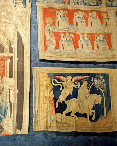 The image shows several large, medieval tapestries with intricate religious and apocalyptic scenes, displayed on a dark blue textured wall inside a museum setting; visible details include Gothic architectural elements, figures in period attire, an
