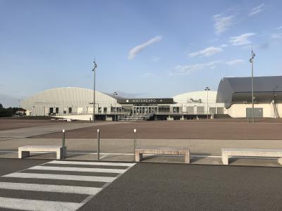 The image shows a large, modern exhibition center complex with distinctive arched white roofs and a central entrance building displaying the sign "AINTEREXPO", set against a blue sky with a paved plaza and benches in the foreground.