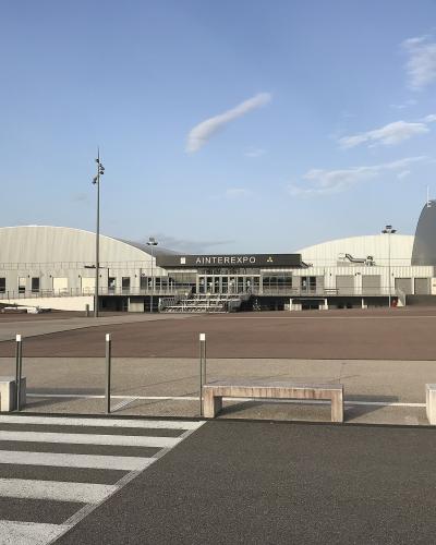 The image shows a large, modern exhibition center complex with distinctive arched white roofs and a central entrance building displaying the sign "AINTEREXPO", set against a blue sky with a paved plaza and benches in the foreground.