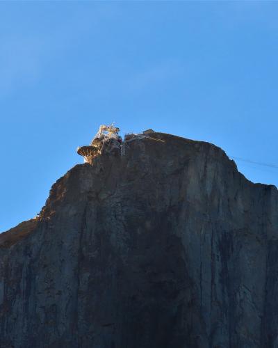 A rocky mountain summit with a cable car station structure and visible cables against a clear blue sky.