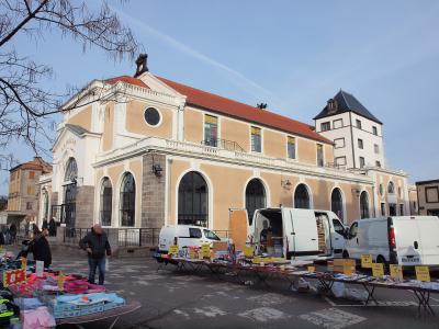The image shows an outdoor market with vendor stalls and white vans parked in front of a large, classical-style building with arched windows, a red-tiled roof, and the inscription "LA HALLE" above the entrance; a tower is visible to the right.