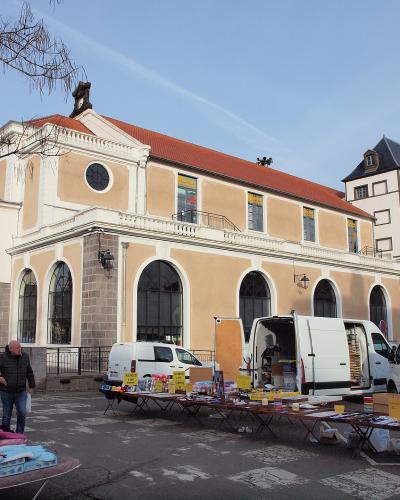 The image shows an outdoor market with vendor stalls and white vans parked in front of a large, classical-style building with arched windows, a red-tiled roof, and the inscription "LA HALLE" above the entrance; a tower is visible to the right.