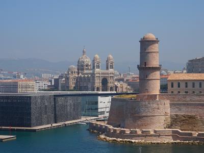 The image shows the MuCEM building with its distinctive dark, perforated concrete facade and glass structure, situated next to the historic Fort Saint-Jean with its prominent round tower, and in the background is the Cathédrale Sainte-Marie-Majeur