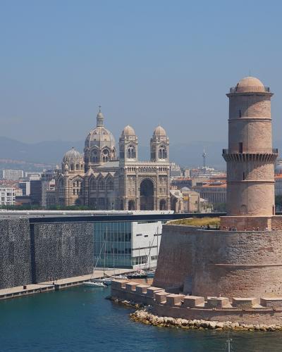 The image shows the MuCEM building with its distinctive dark, perforated concrete facade and glass structure, situated next to the historic Fort Saint-Jean with its prominent round tower, and in the background is the Cathédrale Sainte-Marie-Majeur
