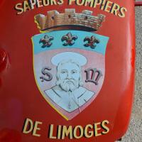 A close-up of a red fire truck’s side panel displaying the emblem and text "SAPEURS-POMPIERS DE LIMOGES," featuring a crowned shield with three fleurs-de-lis and a portrait of Saint Martial, patron saint of Limoges.