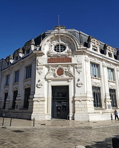 A large, ornate white stone building with "CRÉDIT LYONNAIS" signage on its facade, situated at a street corner with cobblestone pavement and pedestrians nearby.