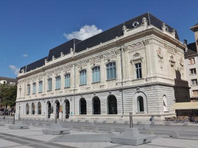 A large, ornate neoclassical building with a dark mansard roof, arched ground-floor windows, and decorative stone carvings, situated in a paved public square under a blue sky.