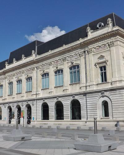 A large, ornate neoclassical building with a dark mansard roof, arched ground-floor windows, and decorative stone carvings, situated in a paved public square under a blue sky.