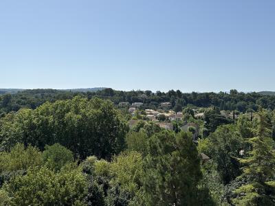 A wide landscape photograph showing a village nestled among dense green trees and rolling hills under a clear blue sky, with no prominent castle structure visible in the foreground or center.