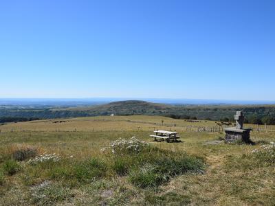 A stone cross on a pedestal in a grassy mountain pasture with a picnic table and distant rolling hills under a clear blue sky.