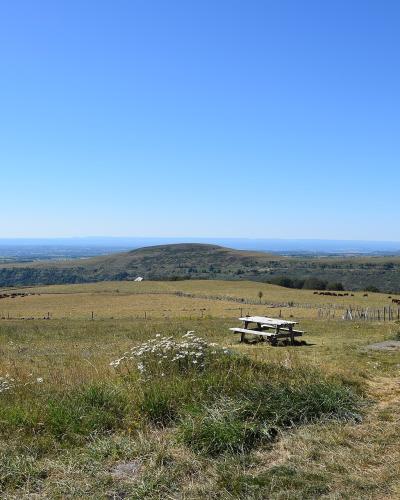 A stone cross on a pedestal in a grassy mountain pasture with a picnic table and distant rolling hills under a clear blue sky.