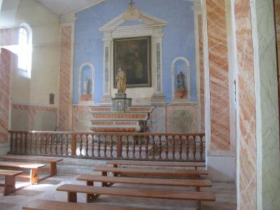 The image shows the interior of a small, modest chapel with a central altar featuring a religious painting and statues, flanked by pinkish marble-like columns and simple wooden pews.