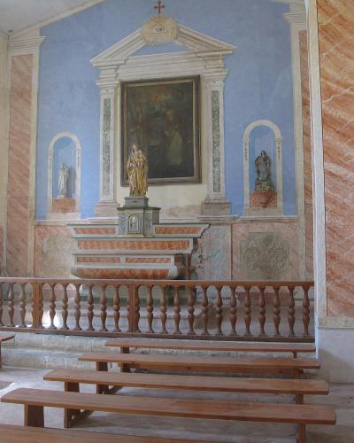 The image shows the interior of a small, modest chapel with a central altar featuring a religious painting and statues, flanked by pinkish marble-like columns and simple wooden pews.
