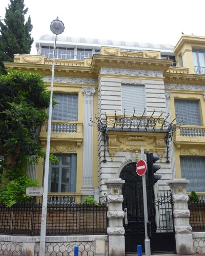 A grand, ornate yellow and white Belle Époque villa with decorative stonework, wrought-iron balconies, and a gated entrance on Rue Constant, as indicated by the street sign.