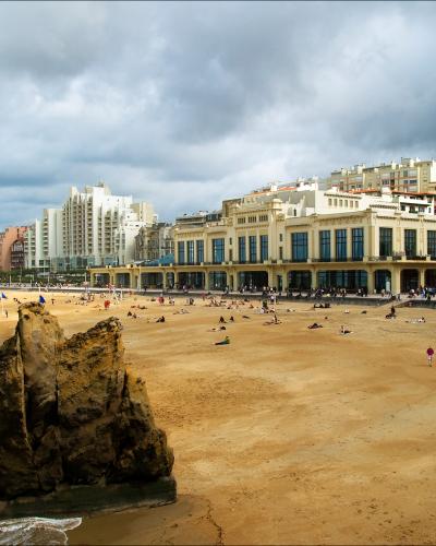A wide sandy beach with a prominent rock formation in the foreground, people scattered across the sand, and a large ornate building labeled "CASINO" along the promenade under a cloudy sky.