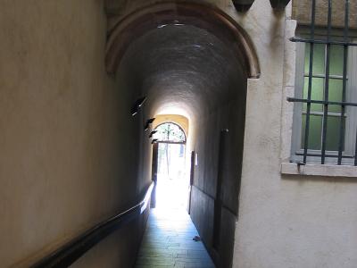 A stone-arched, vaulted passageway with a ramp, leading through a building to an outdoor courtyard, featuring a barred window and modern lighting fixtures.