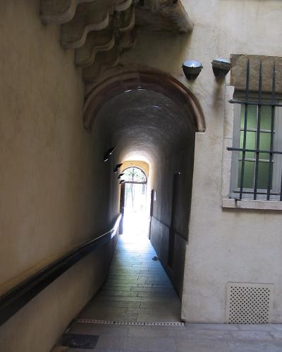 A stone-arched, vaulted passageway with a ramp, leading through a building to an outdoor courtyard, featuring a barred window and modern lighting fixtures.