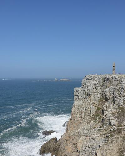 A high-angle view of a rugged, rocky coastal cliff overlooking the sea under a clear blue sky, with a distinctive stone tower (the Tour Vauban) visible on the cliff edge and small figures of people nearby.