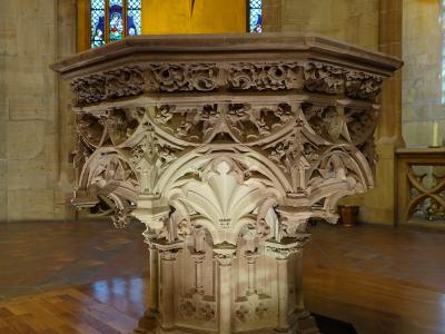 An ornate, octagonal stone baptismal font with intricate Gothic tracery and carvings, situated on a wooden floor inside a church with stained glass windows and vaulted ceilings in the background.