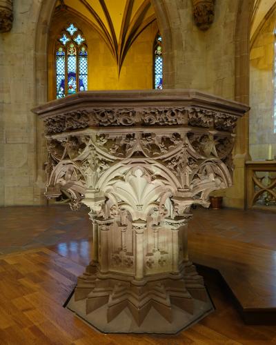 An ornate, octagonal stone baptismal font with intricate Gothic tracery and carvings, situated on a wooden floor inside a church with stained glass windows and vaulted ceilings in the background.