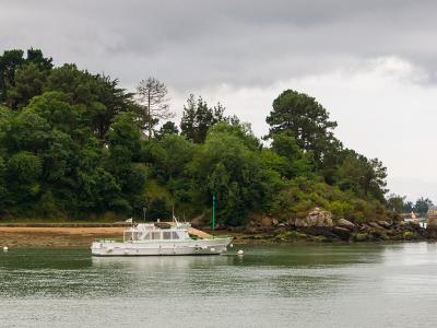 A white passenger boat is moored in calm water near a tree-covered shoreline with rocky outcrops, under an overcast sky; another similar boat is visible further down the channel.
