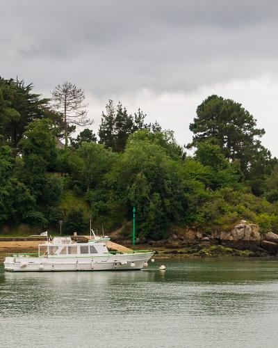 A white passenger boat is moored in calm water near a tree-covered shoreline with rocky outcrops, under an overcast sky; another similar boat is visible further down the channel.