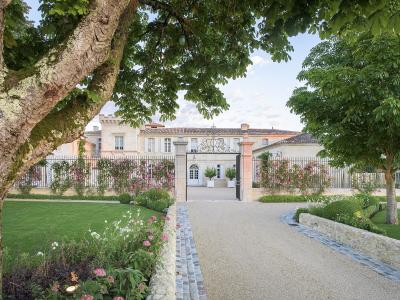 A gravel driveway leads through an open iron gate flanked by stone pillars and climbing pink roses toward a large, light-colored stone château with a tiled roof, framed by mature trees and manicured lawns.