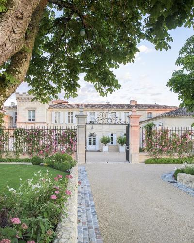 A gravel driveway leads through an open iron gate flanked by stone pillars and climbing pink roses toward a large, light-colored stone château with a tiled roof, framed by mature trees and manicured lawns.