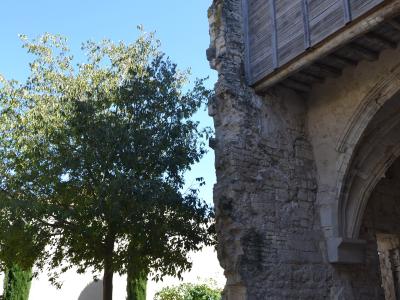 The image shows a stone ruin with a wooden upper structure and an arched opening, adjacent to a gravel courtyard with a tree and bench under a clear blue sky.