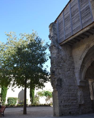 The image shows a stone ruin with a wooden upper structure and an arched opening, adjacent to a gravel courtyard with a tree and bench under a clear blue sky.