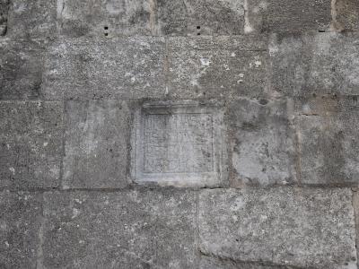 A close-up photograph of a weathered stone masonry wall featuring a carved square stone with Latin inscriptions.