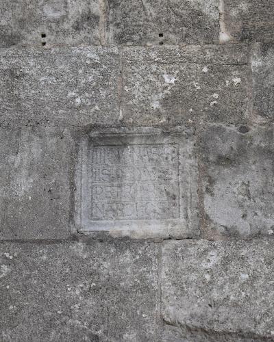 A close-up photograph of a weathered stone masonry wall featuring a carved square stone with Latin inscriptions.