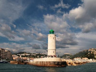 A white cylindrical lighthouse with a green lantern room, situated on a stone pier in a harbor with coastal buildings and hills in the background.