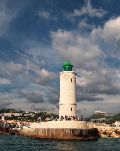 A white cylindrical lighthouse with a green lantern room, situated on a stone pier in a harbor with coastal buildings and hills in the background.
