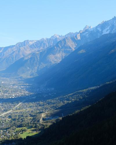 A high-angle landscape photograph showing a valley town nestled between forested slopes, with the prominent jagged peaks of the Mont-Blanc massif visible in the background under a clear blue sky.
