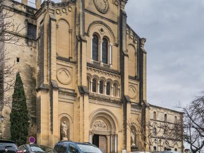 A large, light-colored stone cathedral with Romanesque and Gothic architectural features, including a prominent central tower, arched windows, a statue in a niche, and parked cars in the foreground under an overcast sky.