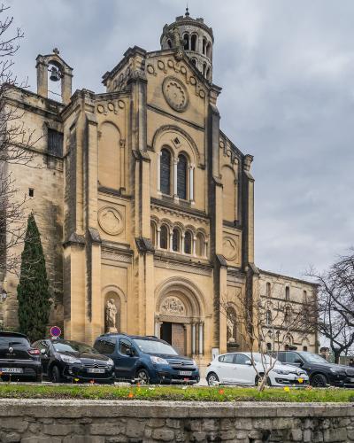 A large, light-colored stone cathedral with Romanesque and Gothic architectural features, including a prominent central tower, arched windows, a statue in a niche, and parked cars in the foreground under an overcast sky.