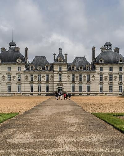 A large, symmetrical French château with a central pavilion and two prominent round towers topped with domed roofs, set behind a gravel courtyard and formal gardens under a cloudy sky.