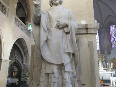 A white marble statue of a priest inside a cathedral, with a plaque at the base reading "CENTENAIRE DE L'ORDINATION SACERDOTALE DE S. J.M. VIANNEY CURE D'ARS A GRENOBLE" and a sign to the left mentioning "Vieux Temple à Grenoble".