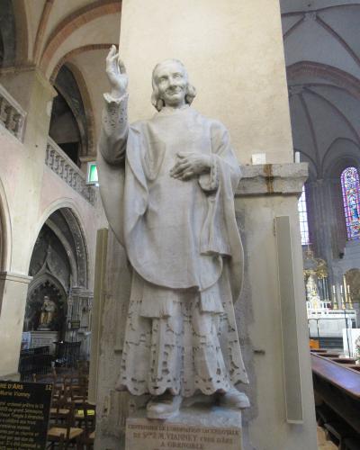 A white marble statue of a priest inside a cathedral, with a plaque at the base reading "CENTENAIRE DE L'ORDINATION SACERDOTALE DE S. J.M. VIANNEY CURE D'ARS A GRENOBLE" and a sign to the left mentioning "Vieux Temple à Grenoble".